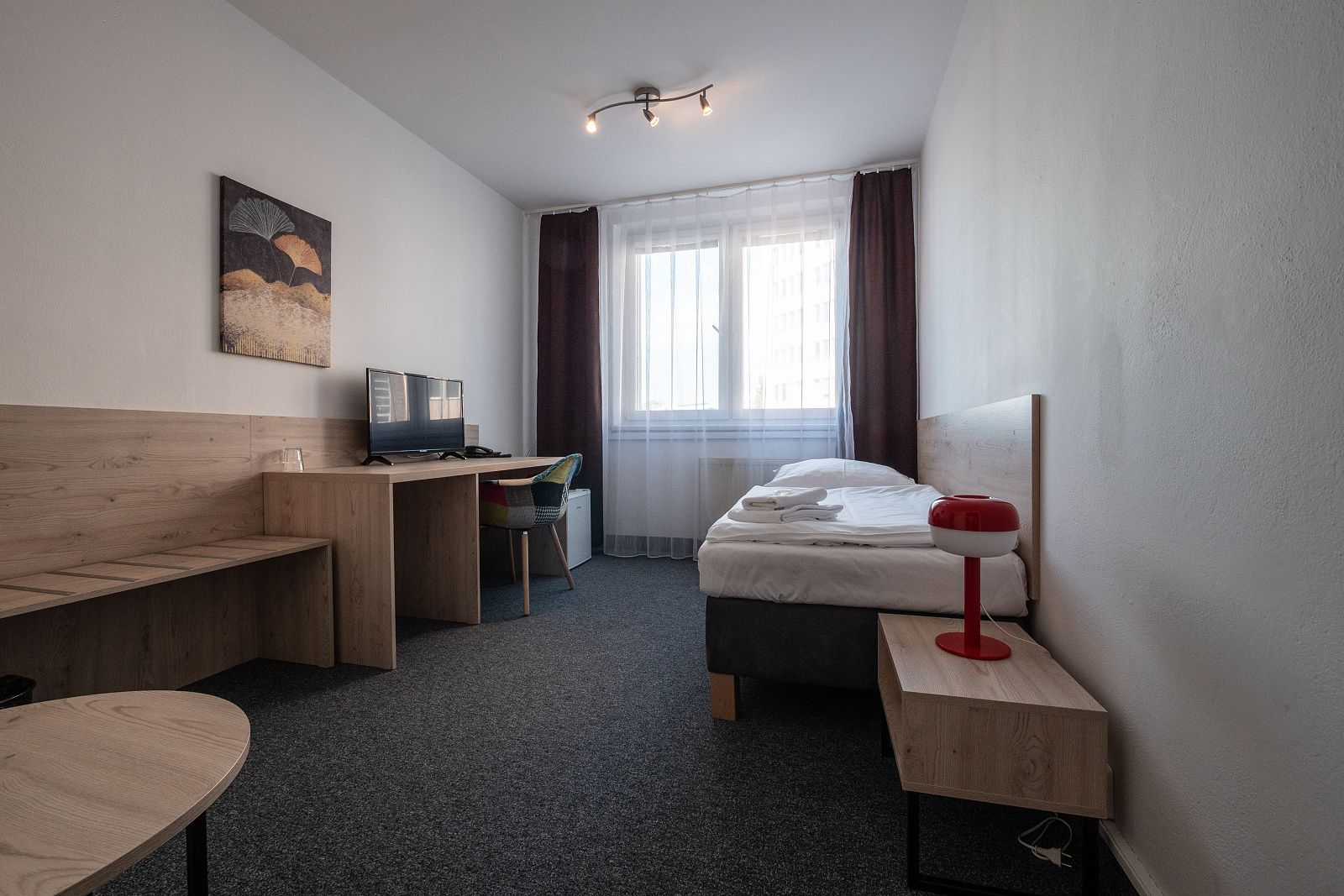 Elegantně zařízený jednolůžkový pokoj s psacím stolem, televizí a červenou lampičkou na stolku